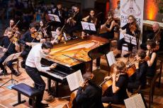 L'image montre un orchestre de jeunes musiciens jouant dans une salle. Au centre se trouve un piano à queue, autour duquel sont disposés des violonistes, violoncellistes et un pianiste.