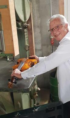 L'image montre un homme âgé souriant, portant des lunettes et une chemise blanche, qui tient un violon.