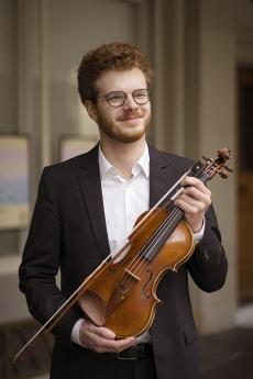 Jeune homme souriant portant des lunettes et une barbe rousse, vêtu d'une chemise blanche et d'une veste noir.