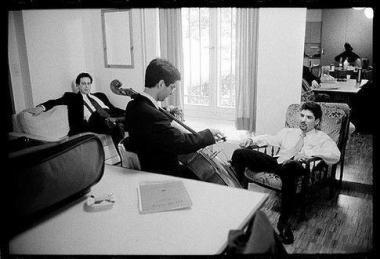 Photo noir et blanc d'un homme qui joue d'un instrument devant deux autres hommes assis dans des fauteilles